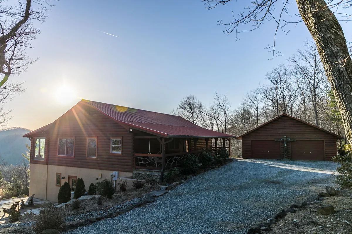 This image features the log cabin structure with its distinctive red metal roof, nestled among mature trees and set along a gravel driveway. The natural surroundings suggest immediate access to outdoor adventures and blend seamlessly into the mountain landscape.