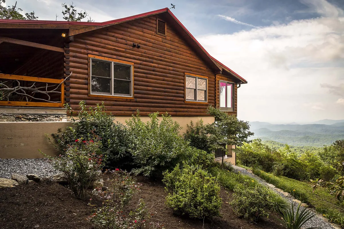A striking exterior view of Alpine Vista's log cabin architecture set against a backdrop of mountains and lush gardens. The porch, visible in the foreground, offers a welcoming and scenic space for families and pets to enjoy the outdoors together.