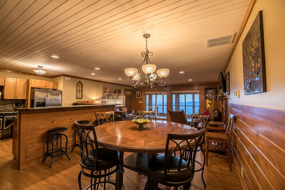 A cozy round wooden dining table sits beneath a chandelier, surrounded by large windows that provide a glimpse of the natural scenery outside. The inviting space is ideal for gathering after outdoor adventures, sharing meals, or casual conversations, with the outdoors always nearby.