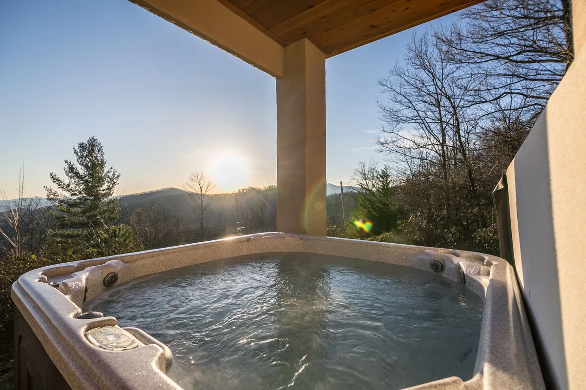 This image captures the exterior hot tub, nestled among trees and overlooking vast mountain vistas as the sun sets in the background. The scene exudes serenity, inviting guests to relax in luxury while surrounded by natural beauty.