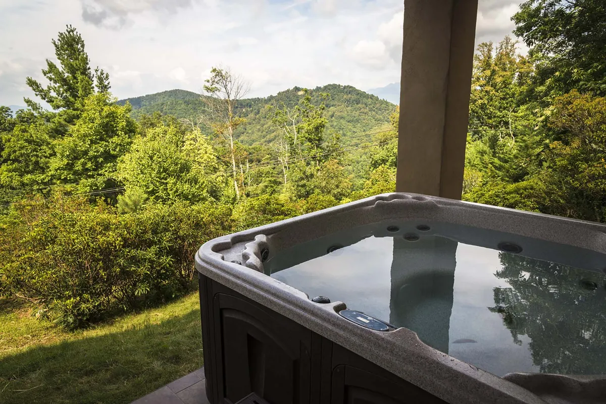 This exterior image features a bubbling hot tub positioned on a deck with a sweeping panoramic view of the mountains and lush greenery beyond. The tranquil setting is perfect for unwinding while soaking in the natural beauty, with sunset and stargazing opportunities evident from the open sky.