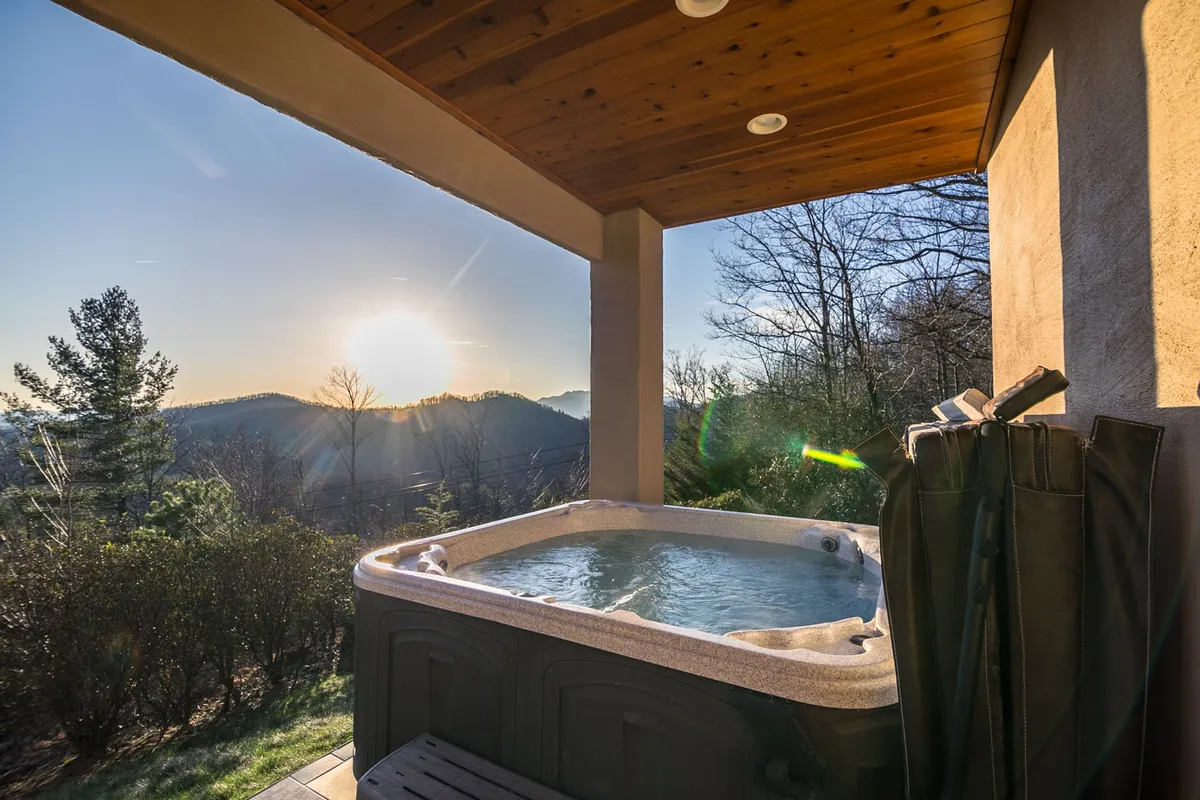The hot tub sits at the edge of the property, offering an unobstructed mountain view and a vibrant sunset sky. The scene evokes relaxation after a day of adventure, with the hot tub as a luxe amenity and the mountain panorama as a backdrop.