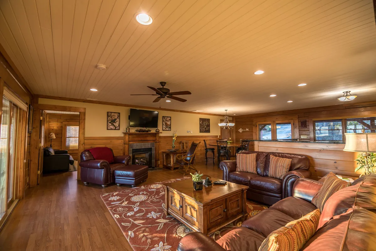 A welcoming view of the great room with leather sofas clustered around a coffee table, a glowing fireplace, and an adjacent dining area. Large windows fill the space with natural light and offer a glimpse of the mountains outside, creating an ideal spot for gatherings and relaxation.