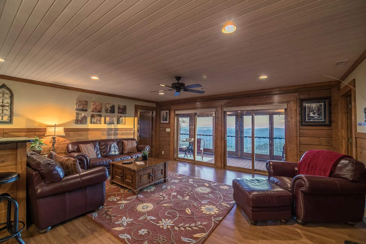 A cozy den area furnished with leather sofas, a coffee table, and a rug, framed by sliding glass doors that allow natural light to fill the space. The setting is ideal for reading, relaxing, or enjoying movie nights.