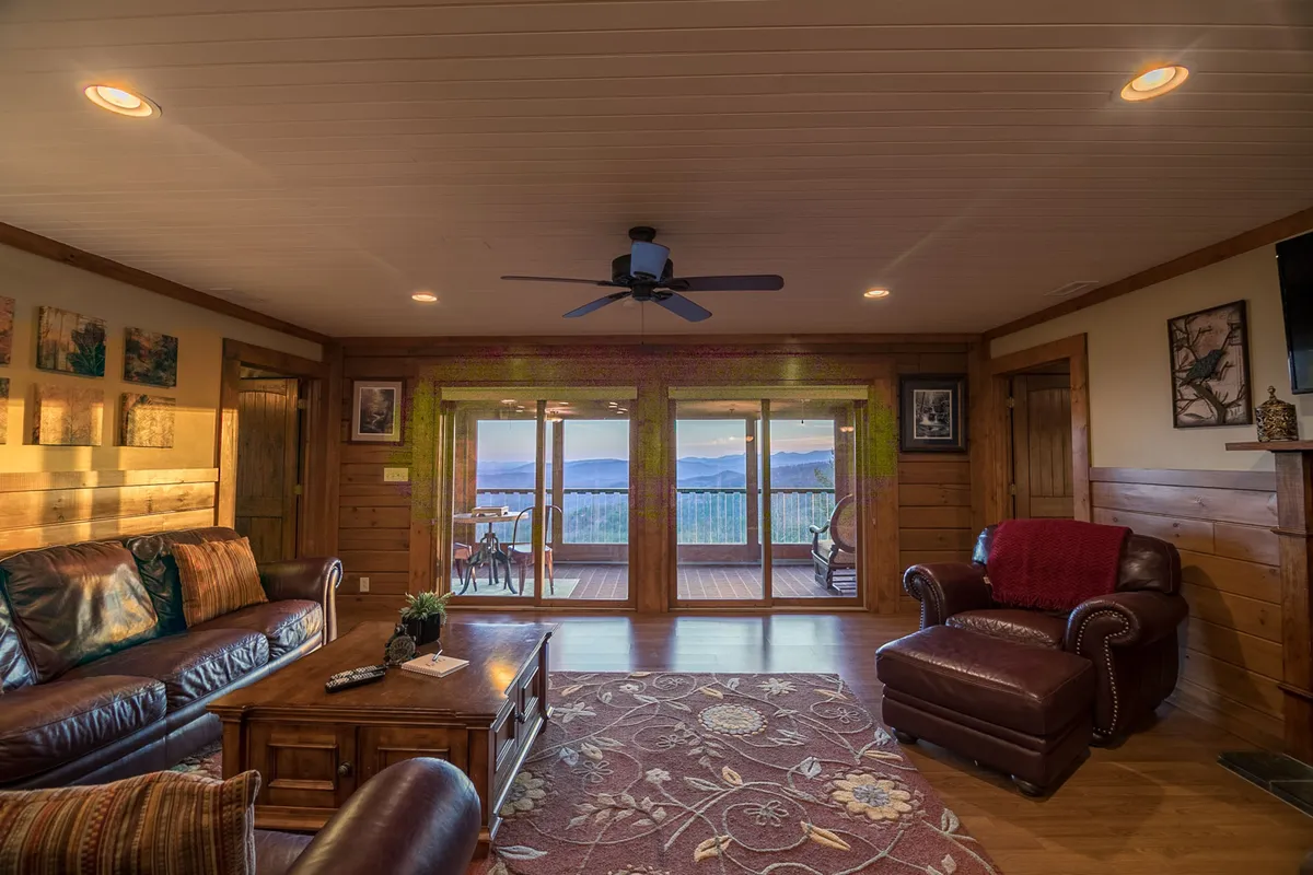 This image captures a spacious great room featuring a comfortable leather sofa, a wooden coffee table, and large windows showcasing breathtaking mountain views. The decor is warm and inviting, with a decorative rug and ceiling fan adding to the cozy ambiance. Sunlight pours into the space, emphasizing the connection to the Blue Ridge vistas beyond.