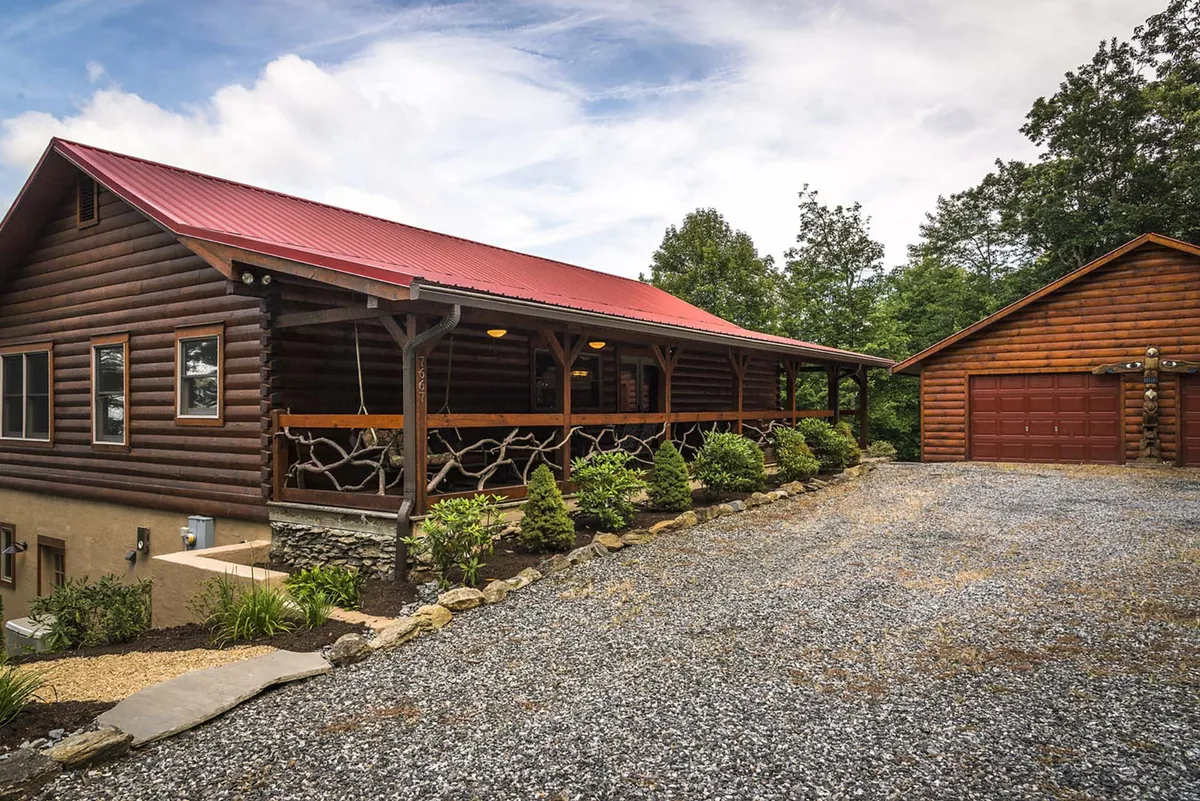 A charming log cabin exterior features a welcoming front porch with a red roof and a landscaped garden, set against a backdrop of lush greenery and mountain scenery. The porch invites guests to gather outdoors, enjoy the fresh air, and connect with the peaceful environment.